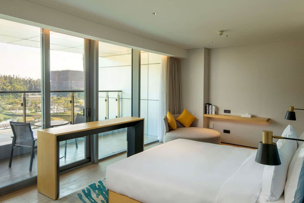 A modern hotel room with a large bed, a desk, a chair, a sofa, and a balcony with a view of the city.