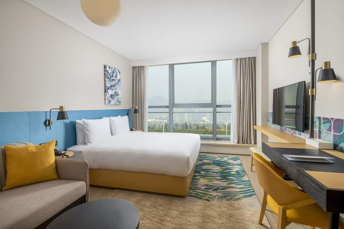 A hotel room with a bed, a desk, a TV, and a large window with a city view.