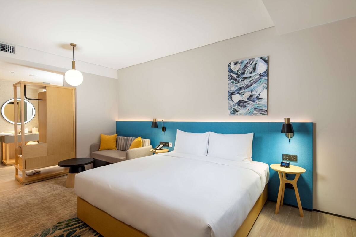A modern hotel room with a large bed, a bedside table, a small stool, a nightstand, and a wall-mounted lamp.