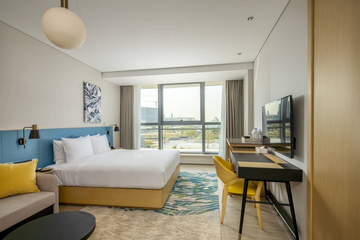 A modern hotel room with a large bed, a desk, a chair, a television, and a view of the city.
