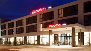 A modern hotel with a prominent "Hampton" sign, large windows, and a well-lit entrance area.