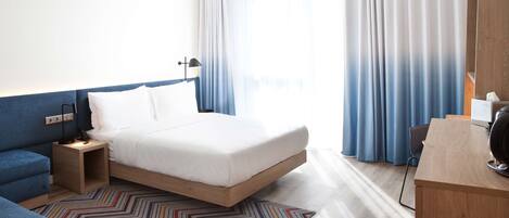 A hotel room with a bed, a desk, a chair, a nightstand, and a patterned rug.
