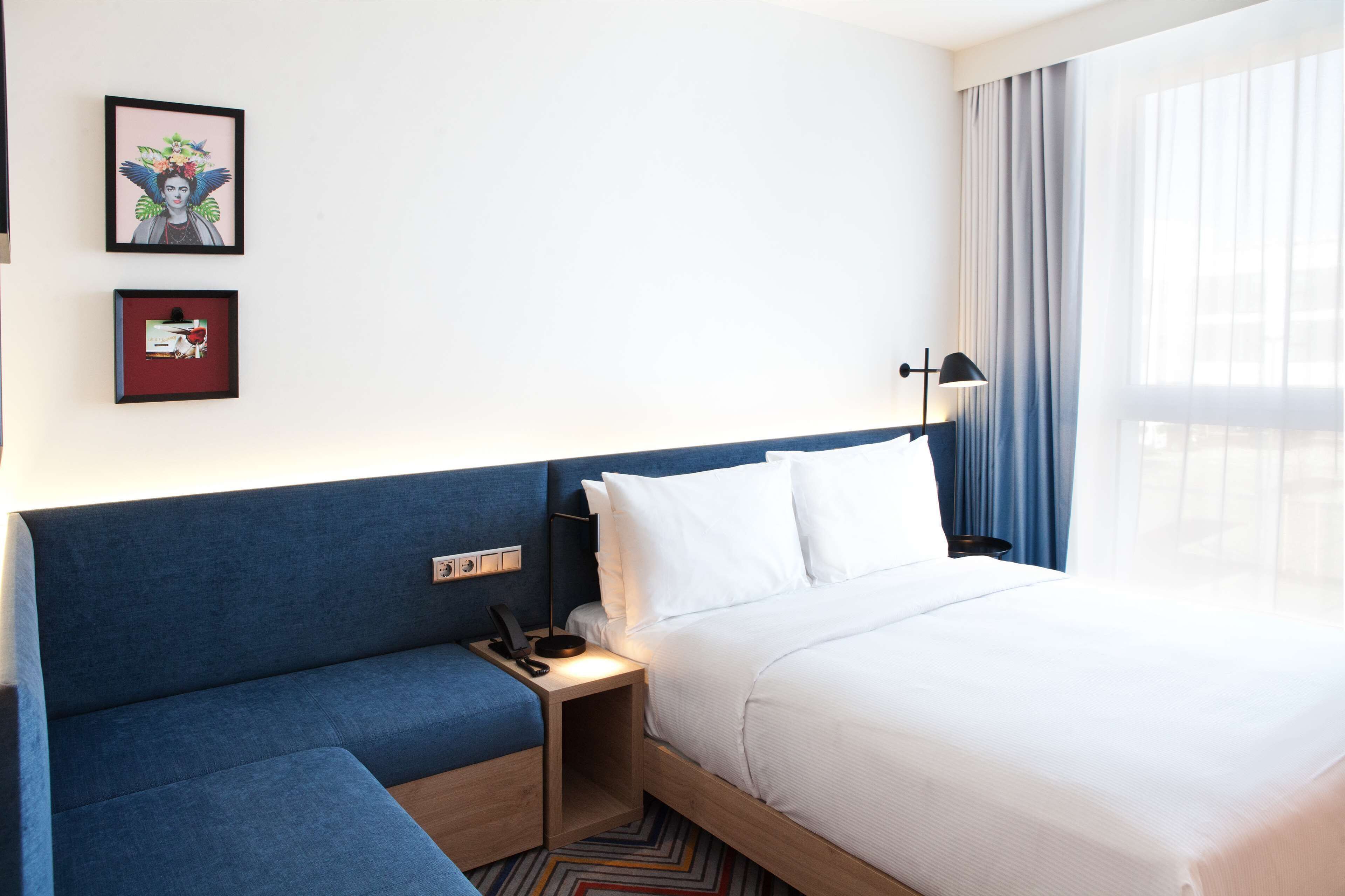 A hotel room with a bed, a blue sofa, a bedside table with a lamp, and two framed artworks on the wall.