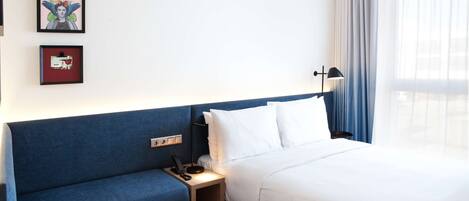 A hotel room with a bed, a blue sofa, a bedside table with a lamp, and two framed artworks on the wall.
