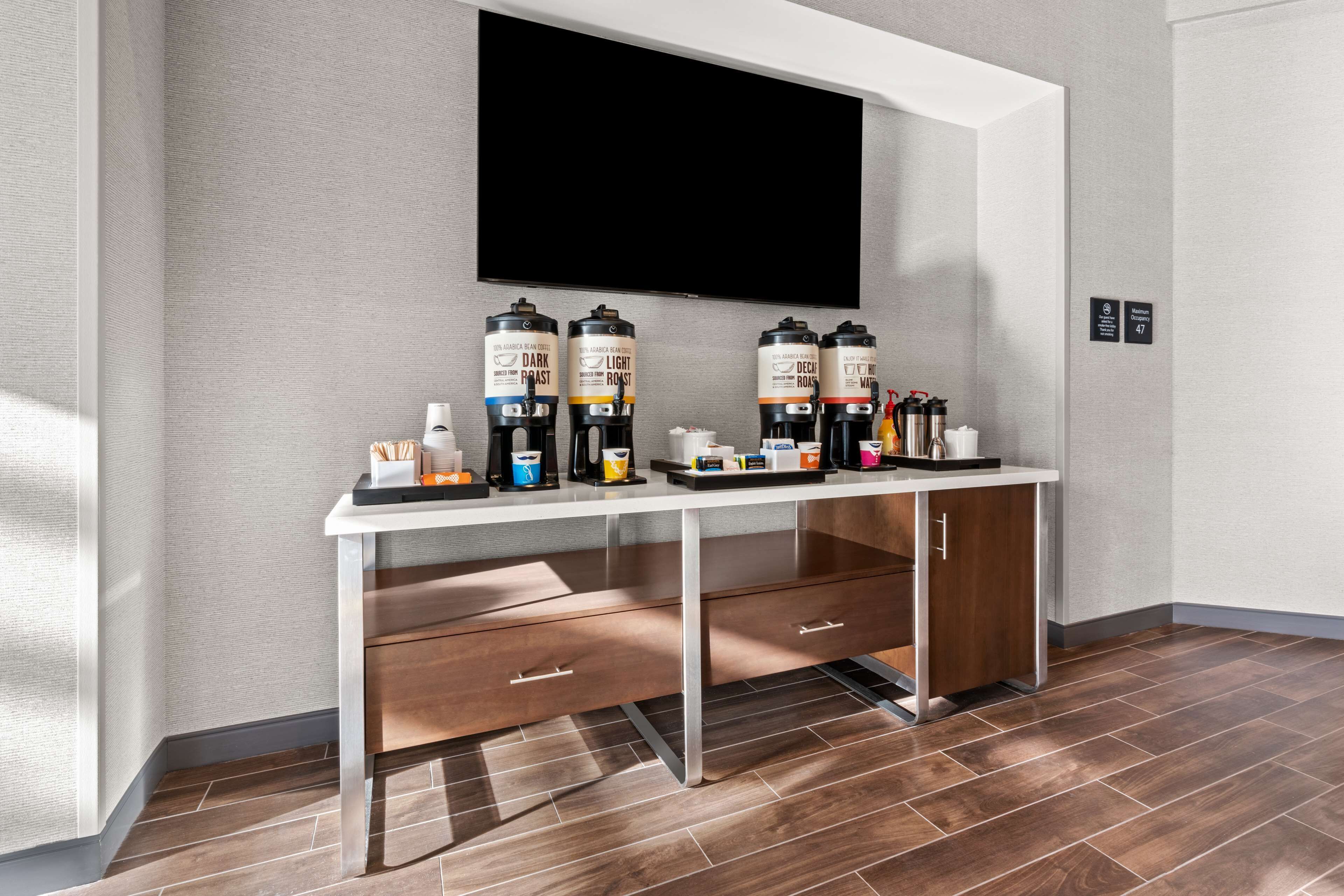 Ett modernt hotellrum med en platt-TV som är monterad på väggen, en kaffestation med flera kaffedispensrar och ett träskåp med lådor.