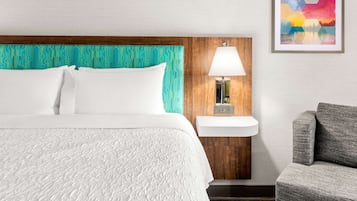 A hotel room with a bed, a nightstand with a lamp, a gray armchair, and a wall-mounted shelf.
