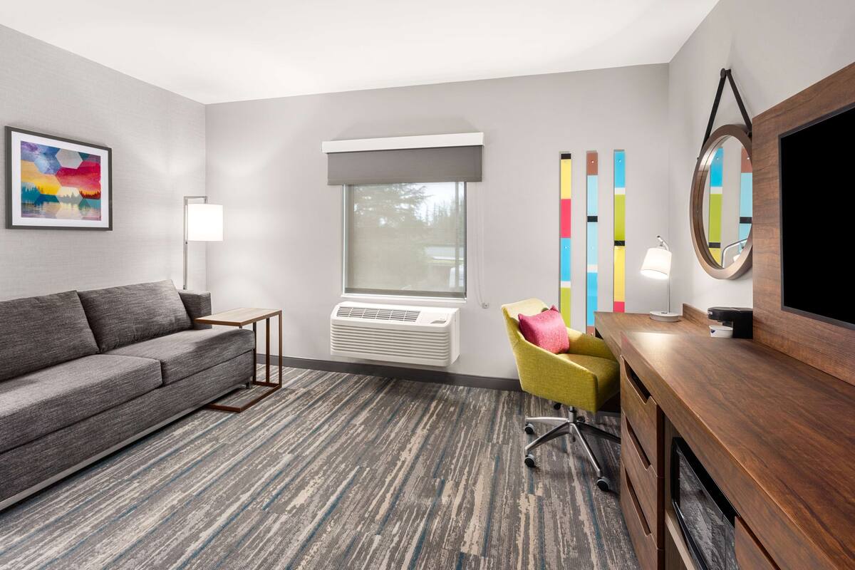 Ett modernt hotellrum med en grå soffa, en gul fåtölj, ett skrivbord i trä, en platt-TV, ett stort fönster med en grå persienn, en golvlampa och en färgstark, abstrakt tavla på väggen.
