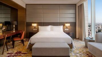 A hotel room with a large bed, a desk with a chair, a small table, and a view of the city.