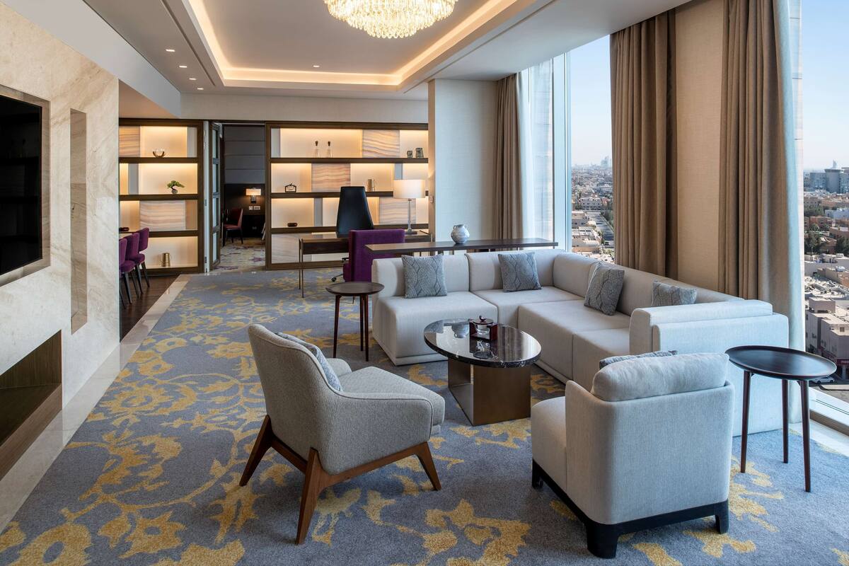 Ein modernes Hotelzimmer mit einer großen Couch, Sesseln, einem Couchtisch und einem Fernseher. Durch große Fenster bietet es einen Blick auf die Stadt.