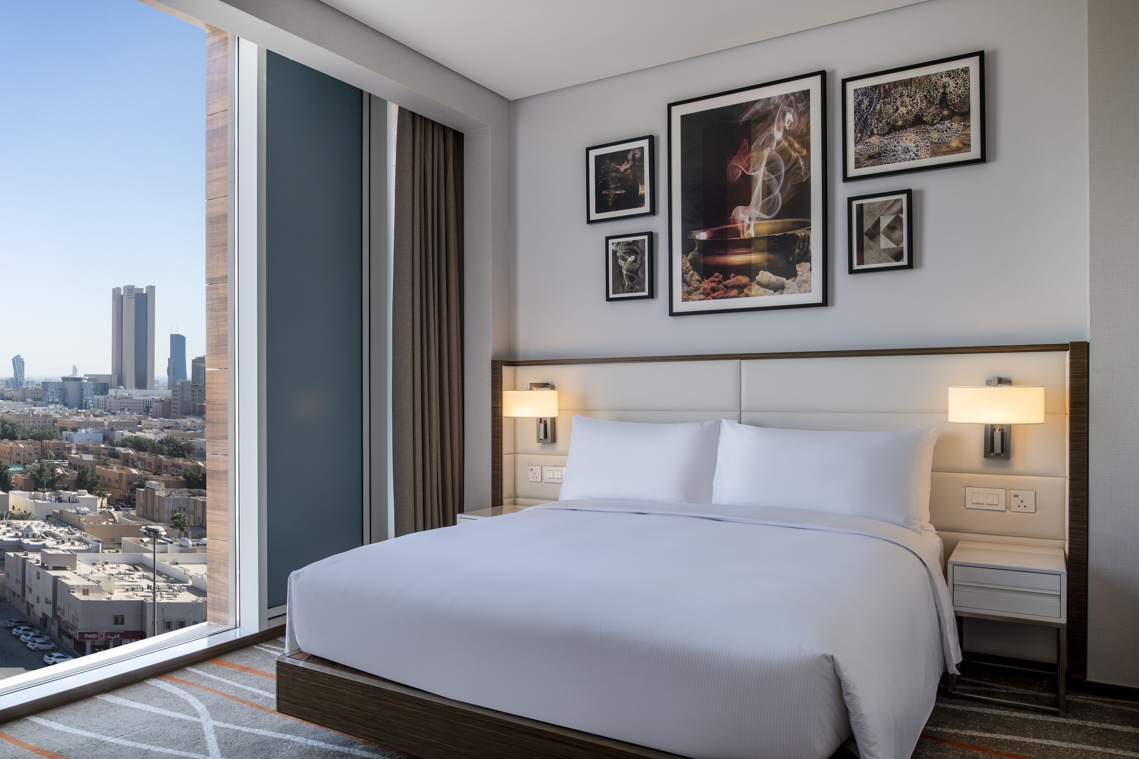 A modern hotel room with a large bed, two bedside tables, a city view, and framed artwork on the wall.