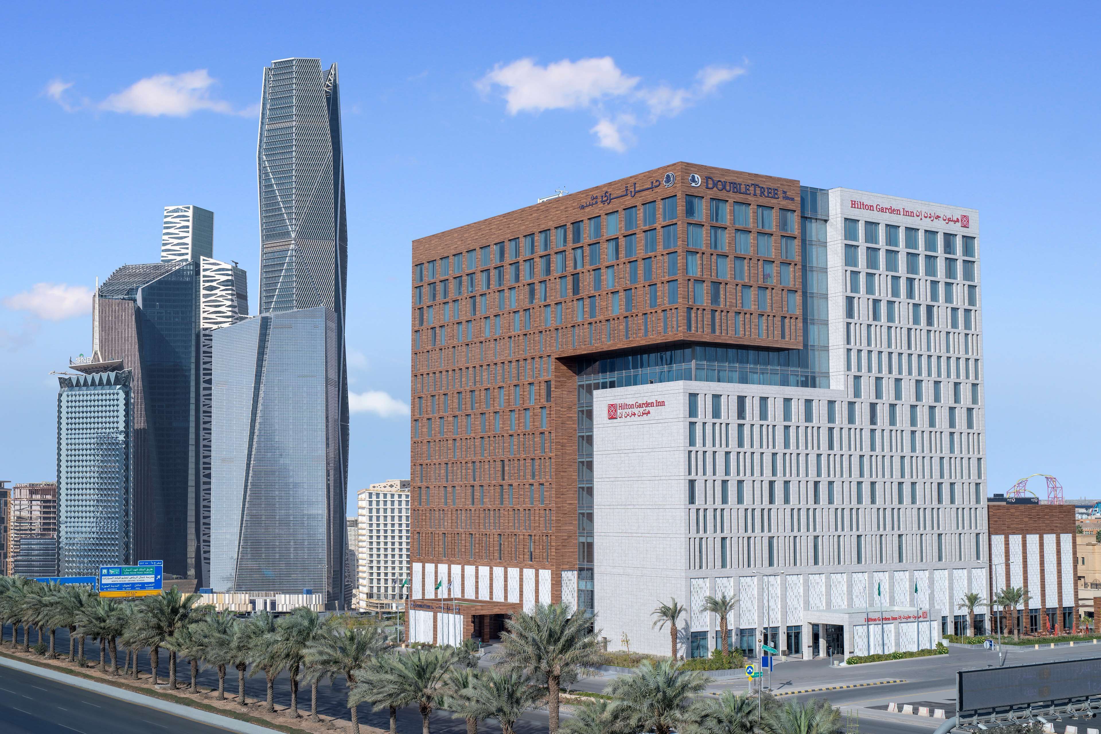 Un hotel moderno con un llamativo letrero de Hilton Garden Inn, rodeado de edificios altos y palmeras.