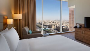 A hotel room with a large bed, a chair, a desk, and a view of a cityscape through large windows. - Hilton Garden Inn Riyadh Financial District (Riyadh)