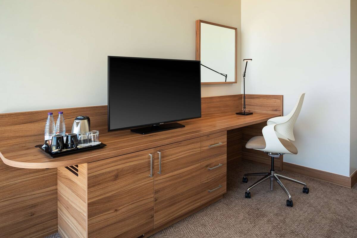 Una habitación de hotel moderna con un escritorio de madera, un televisor de pantalla plana, una silla blanca y una bandeja con vasos y una cafetera.