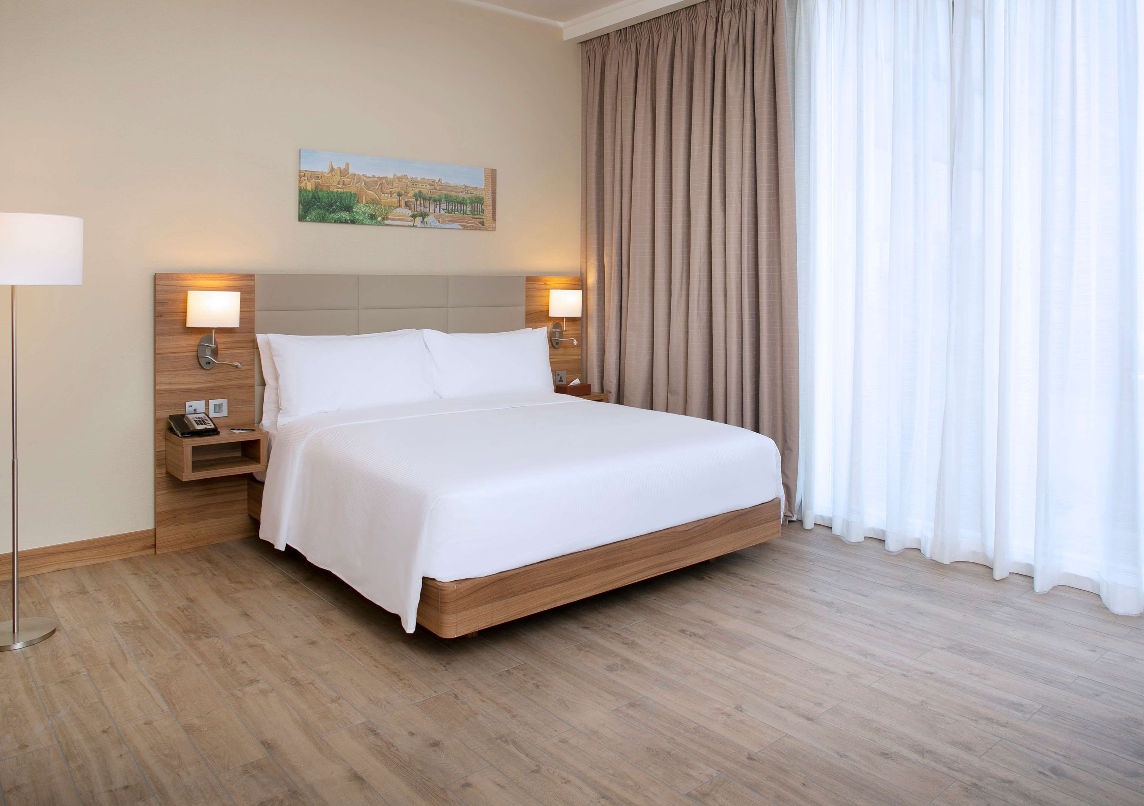 A hotel room with a large bed, wooden headboard, bedside lamps, a nightstand, and a painting on the wall.