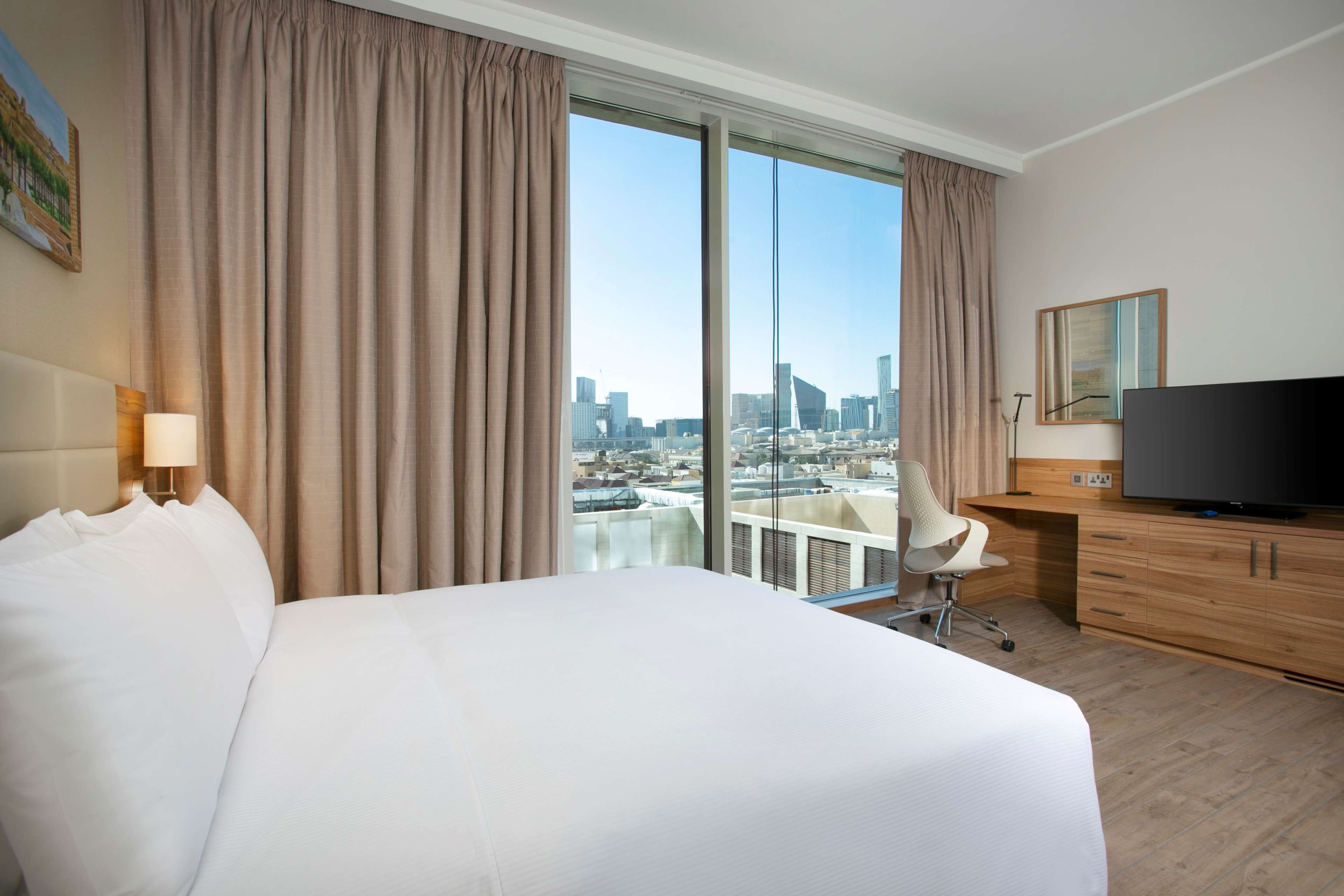 Habitación de hotel con una cama grande, un escritorio, un televisor y vistas a la ciudad a través de amplias ventanas.