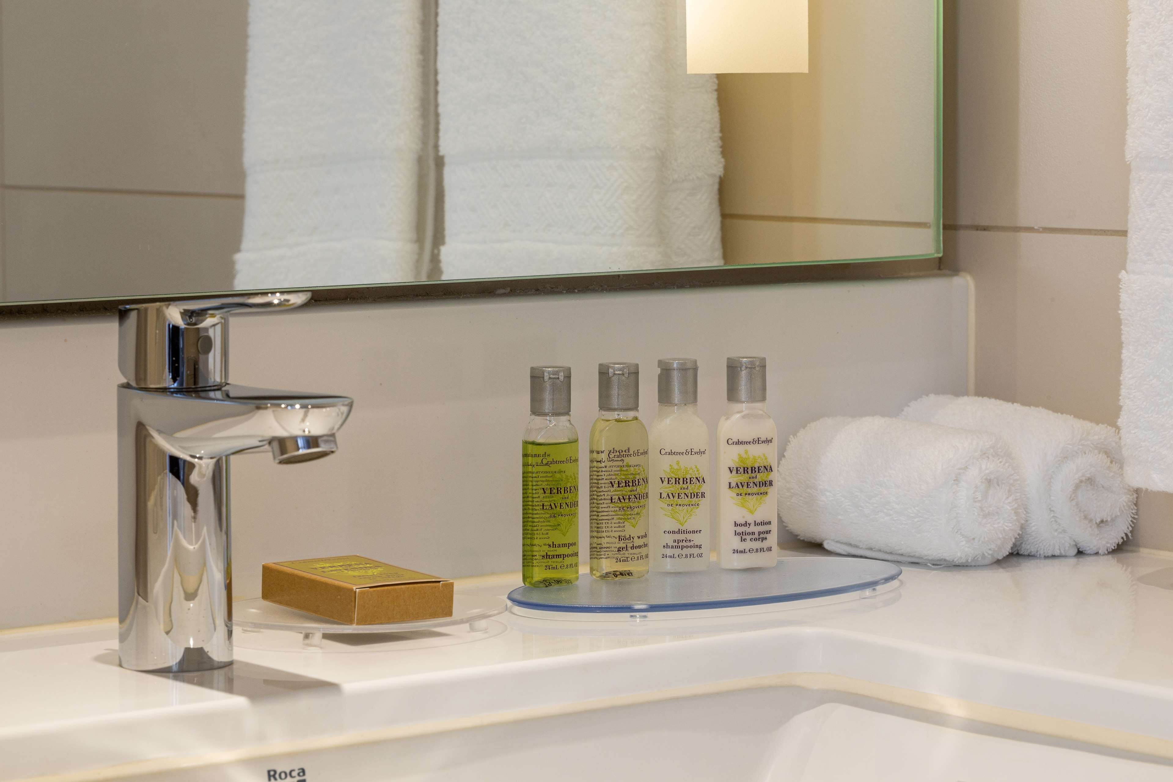 A bathroom with a chrome faucet, a mirror, and a shelf with toiletries including bottles of Verbenia Lavender body lotion and shampoo.