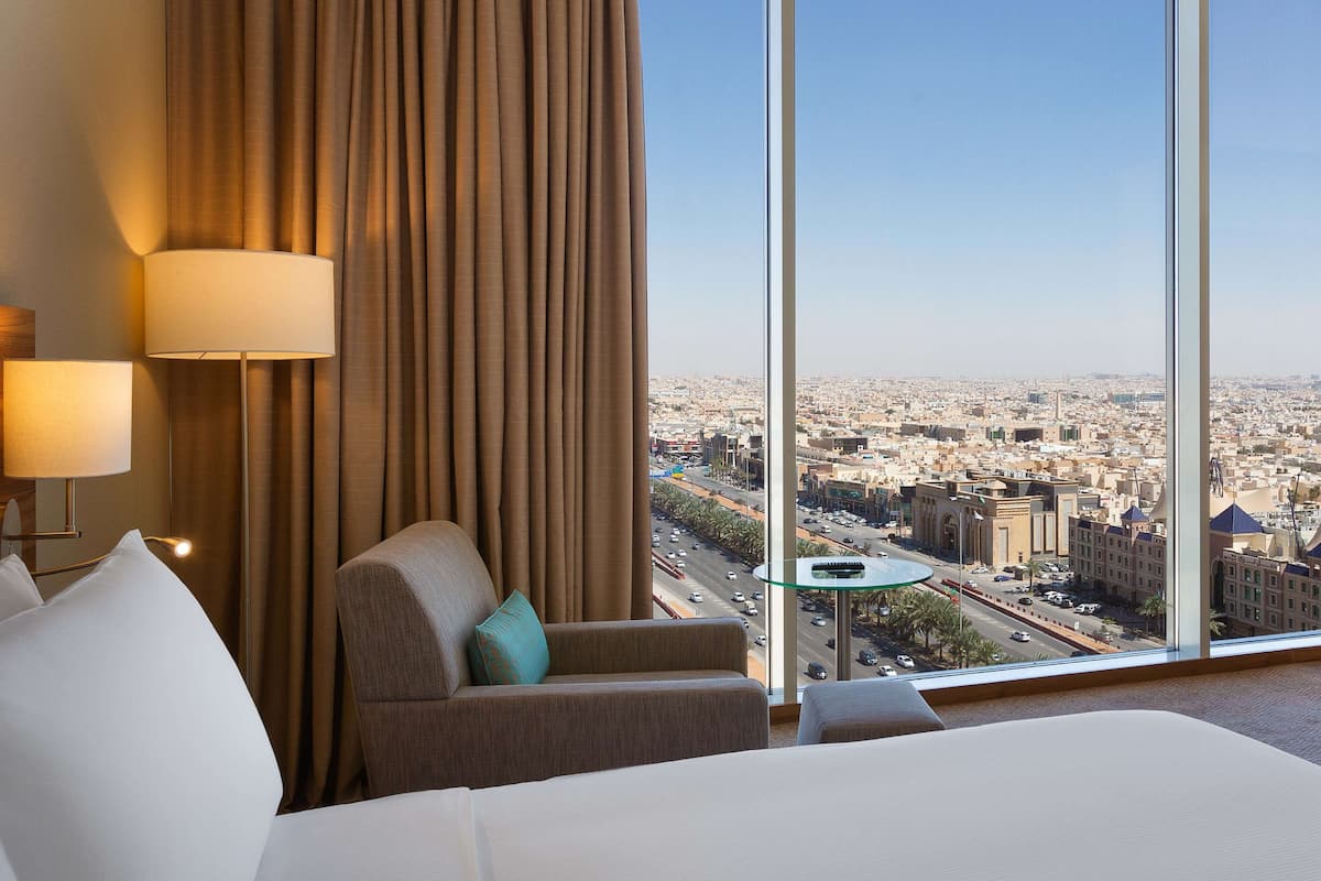 Habitación de hotel con un ventanal amplio que ofrece vistas a la ciudad, una cama, un sillón y una mesita.