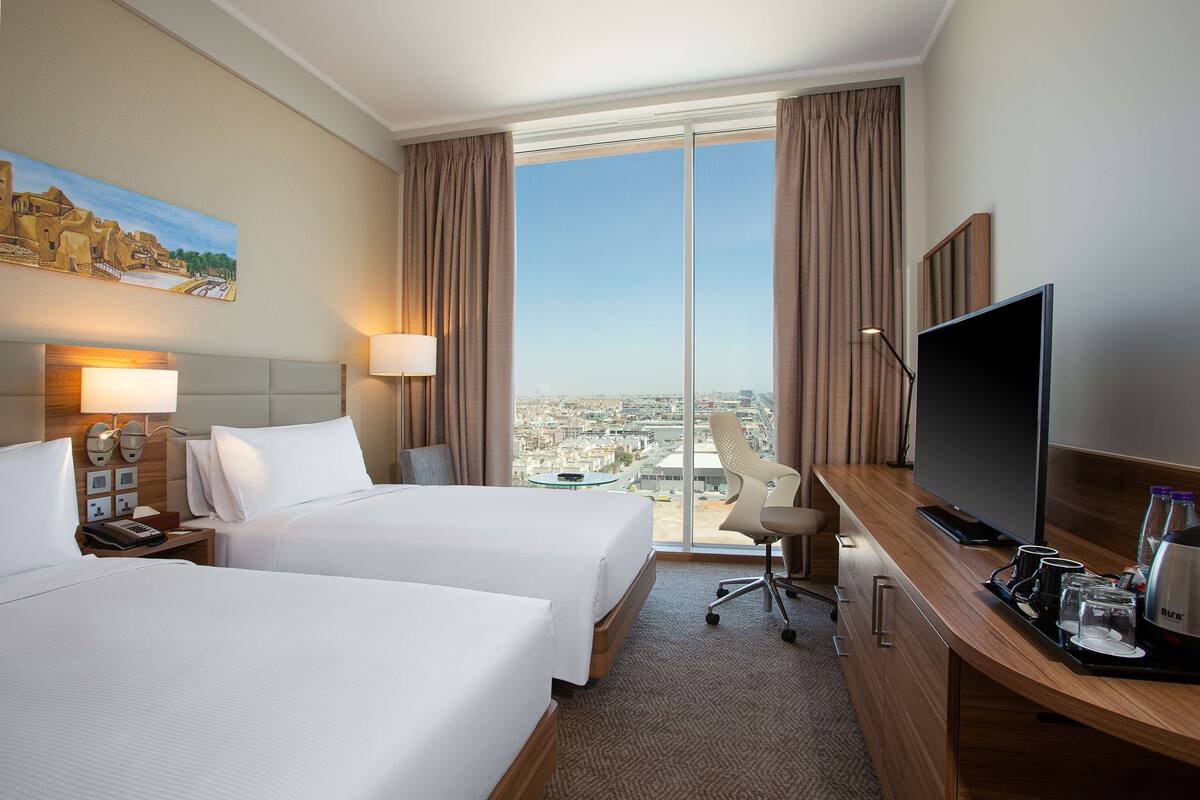 Habitación de hotel con dos camas, un escritorio con televisor y un ventanal con vistas a la ciudad.