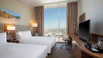 A hotel room with two beds, a desk with a TV, and a large window offering a city view.