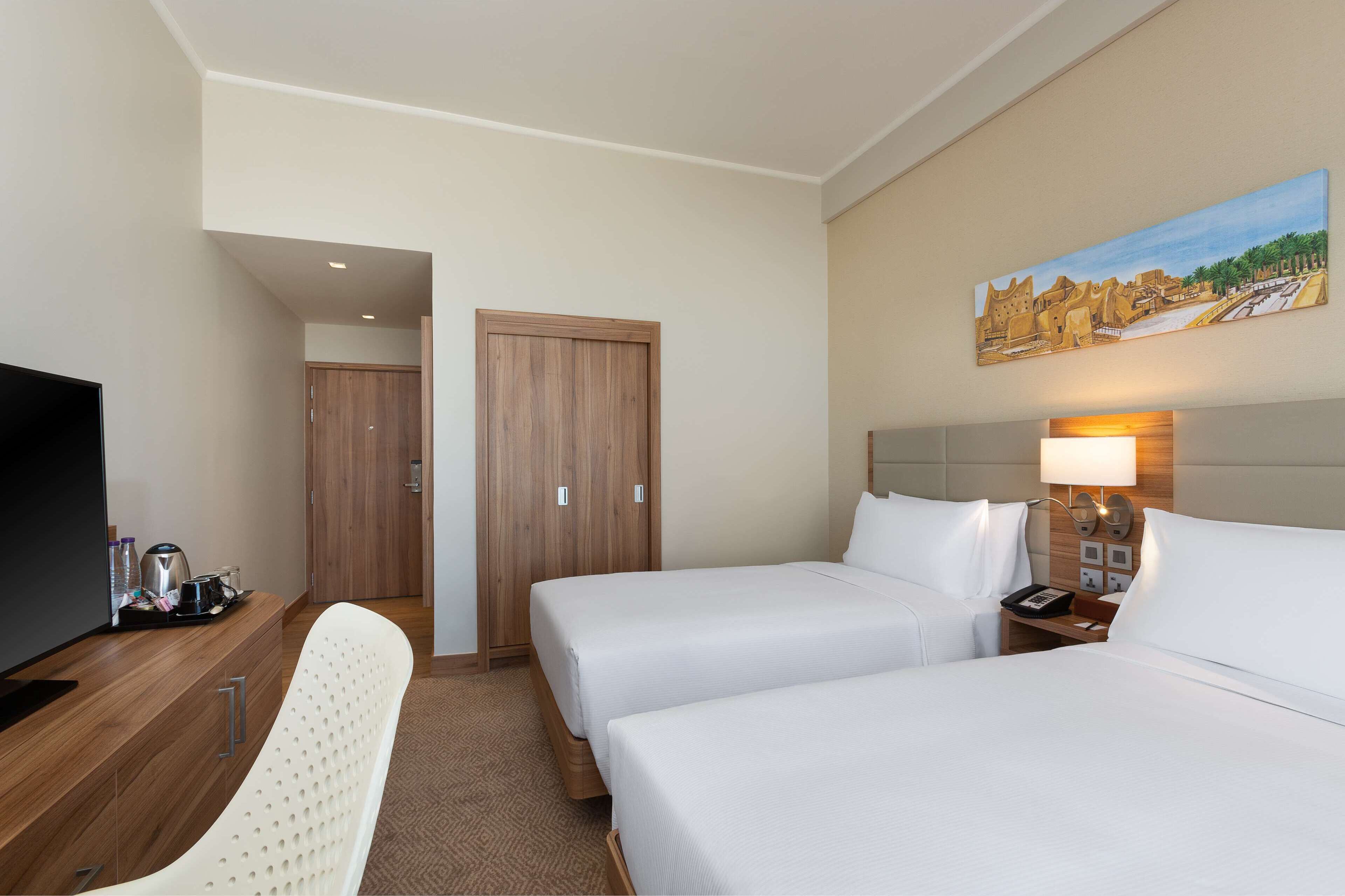 A hotel room with two beds, a desk, a TV, and a painting on the wall.