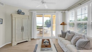 Un salon avec un canapé blanc, une table basse et un meuble. Une porte-fenêtre coulissante donne accès à une terrasse extérieure.