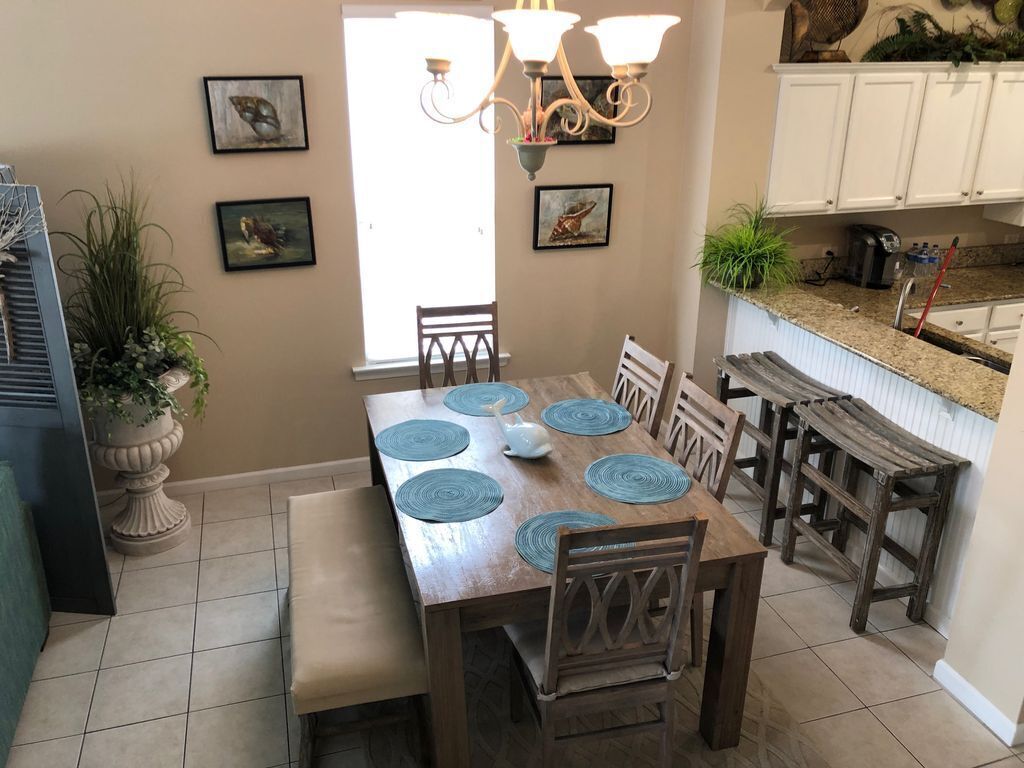 A dining area with a wooden table, blue placemats, and a chandelier. There are framed pictures on the wall and a potted plant on the floor.