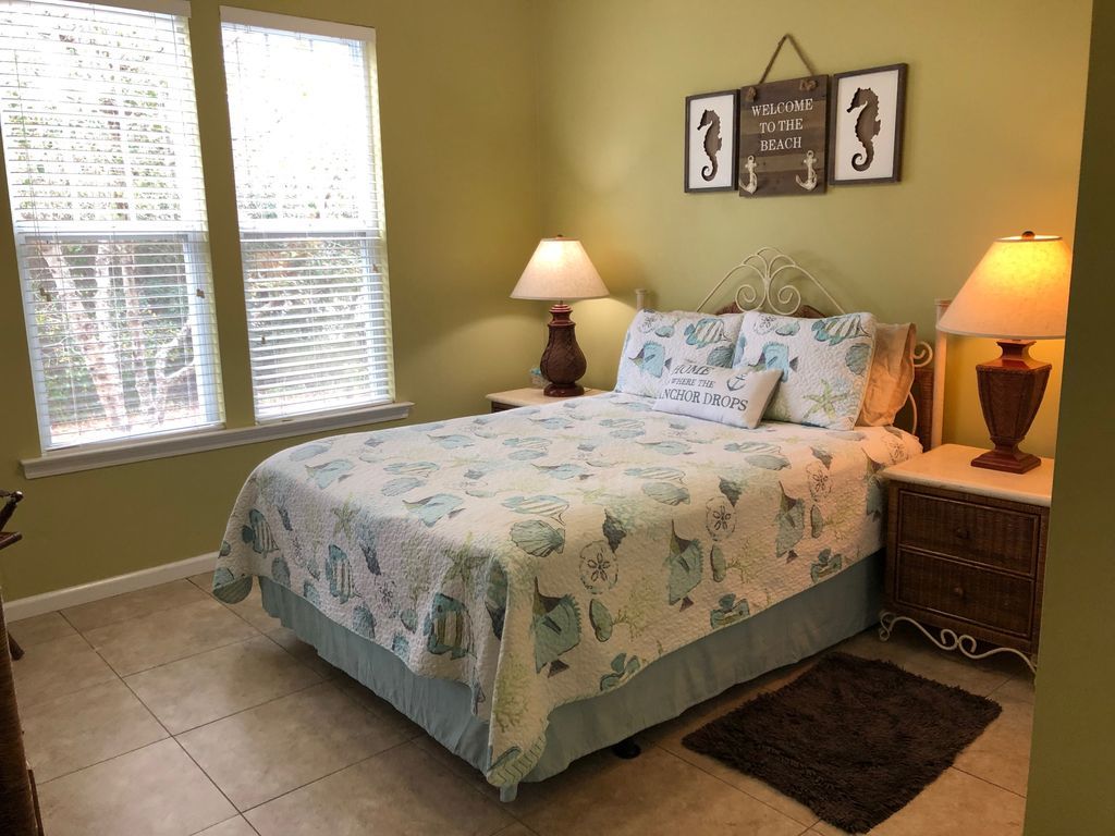 Quarto com uma cama, duas luminárias de cabeceira, um tapete e uma parede com um letreiro que diz 'Bem-vindo à Praia'.