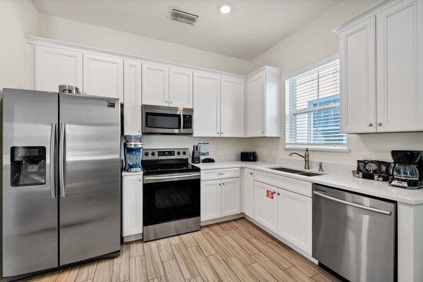 A modern kitchen with white cabinets, stainless steel appliances, a microwave, oven, dishwasher, and a window with blinds.