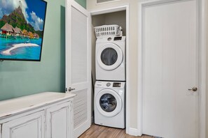 A laundry room with two stacked washing machines, a white door, and a wall-mounted television displaying a tropical scene.