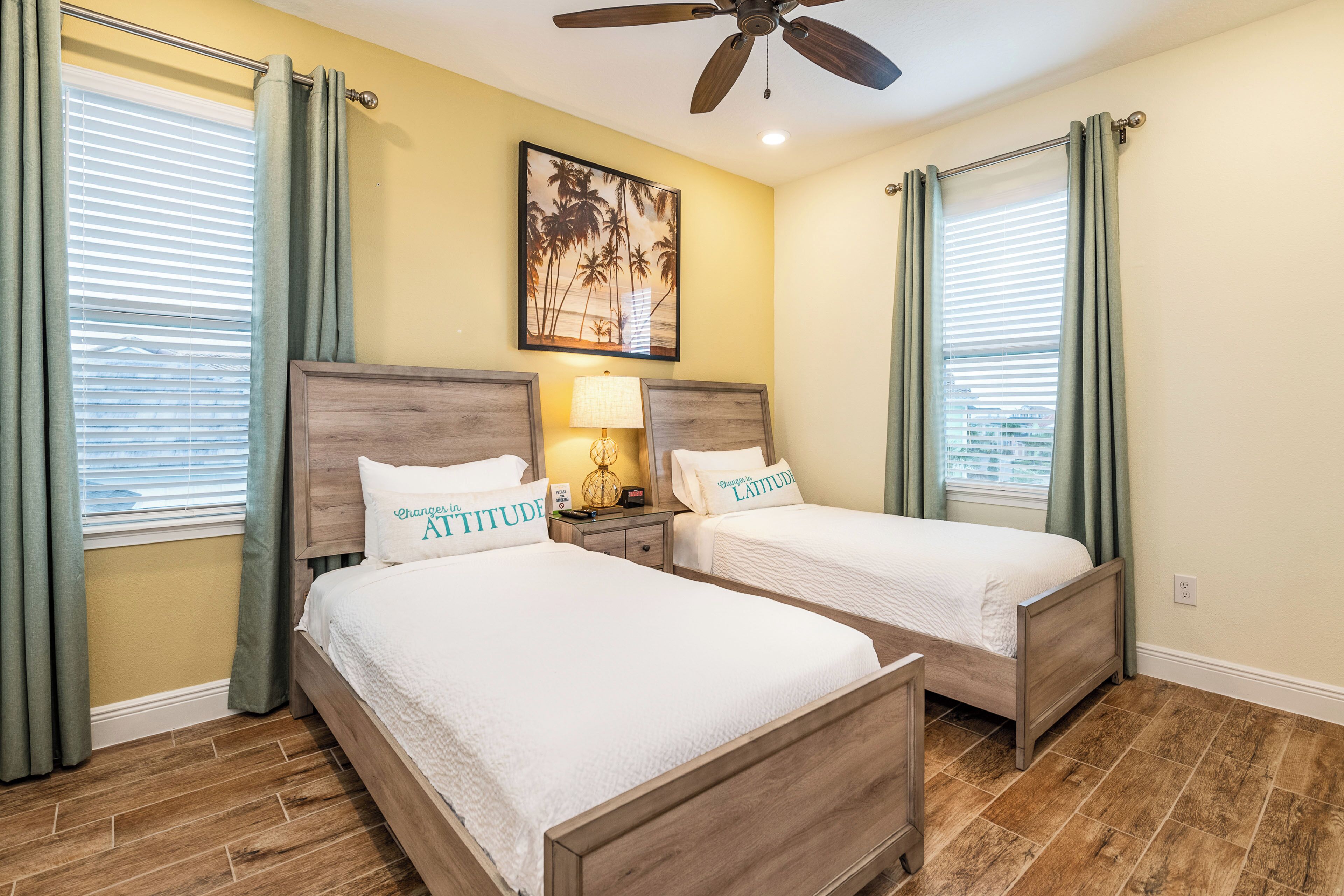 A hotel room with two beds, a ceiling fan, a painting of palm trees, and a window with blinds.
