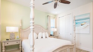 A bedroom with a four-poster bed, a ceiling fan, a nightstand with a lamp, and a painting on the wall.