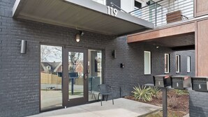 Un edificio moderno con fachada de ladrillo oscuro, número 119, que cuenta con un acceso por puerta de vidrio, un pequeño espacio de asientos al aire libre y buzones.