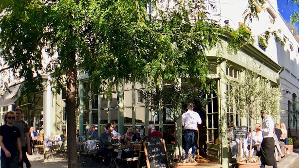 Ein Straßencafé mit Tischen, Stühlen, einer Kreidetafel und einem Gebäude mit dem Schild "The Trans Center".