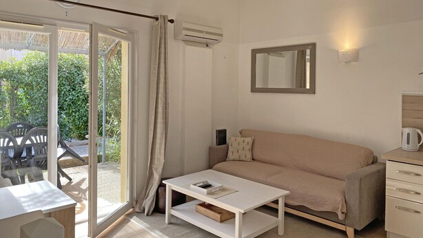 Une pièce comprenant un canapé, une table basse, un bureau et une chaise. Une porte-fenêtre coulissante donne accès à un espace extérieur aménagé avec des chaises et de la verdure.