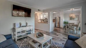A modern living room with a sofa, coffee table, and a TV mounted on the wall. A kitchen is visible in the background with cabinets and a refrigerator. - 443 By the Sea - Gated Myrtle Beach Resort - Near MYR (Myrtle Beach)