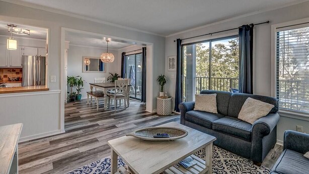 A modern living room with a grey sofa, a coffee table, and a dining area with a chandelier. - 443 By the Sea - Gated Myrtle Beach Resort - Near MYR (Myrtle Beach)
