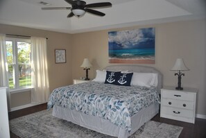 Une chambre à coucher avec un lit, deux tables de chevet, un ventilateur de plafond, une fenêtre avec des rideaux, un tableau représentant une plage et un tableau encadré accroché au mur.