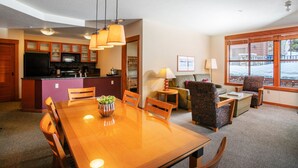 A modern hotel room with a wooden dining table, chairs, a sofa, and a patterned armchair. There is a kitchen area with a refrigerator and microwave. - Spacious Corner VILLAGE Condo 2BD/2BA #2202 w/ Ski-back Trail close to GONDOLA (Mammoth Lakes)