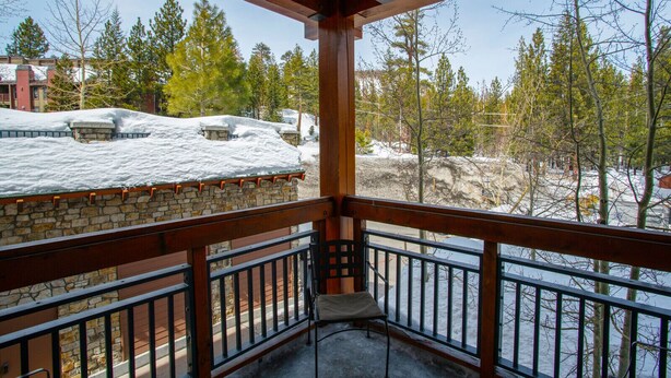 A wooden balcony with a stone wall, overlooking a snowy landscape with trees and buildings. - Spacious Corner VILLAGE Condo 2BD/2BA #2202 w/ Ski-back Trail close to GONDOLA (Mammoth Lakes)