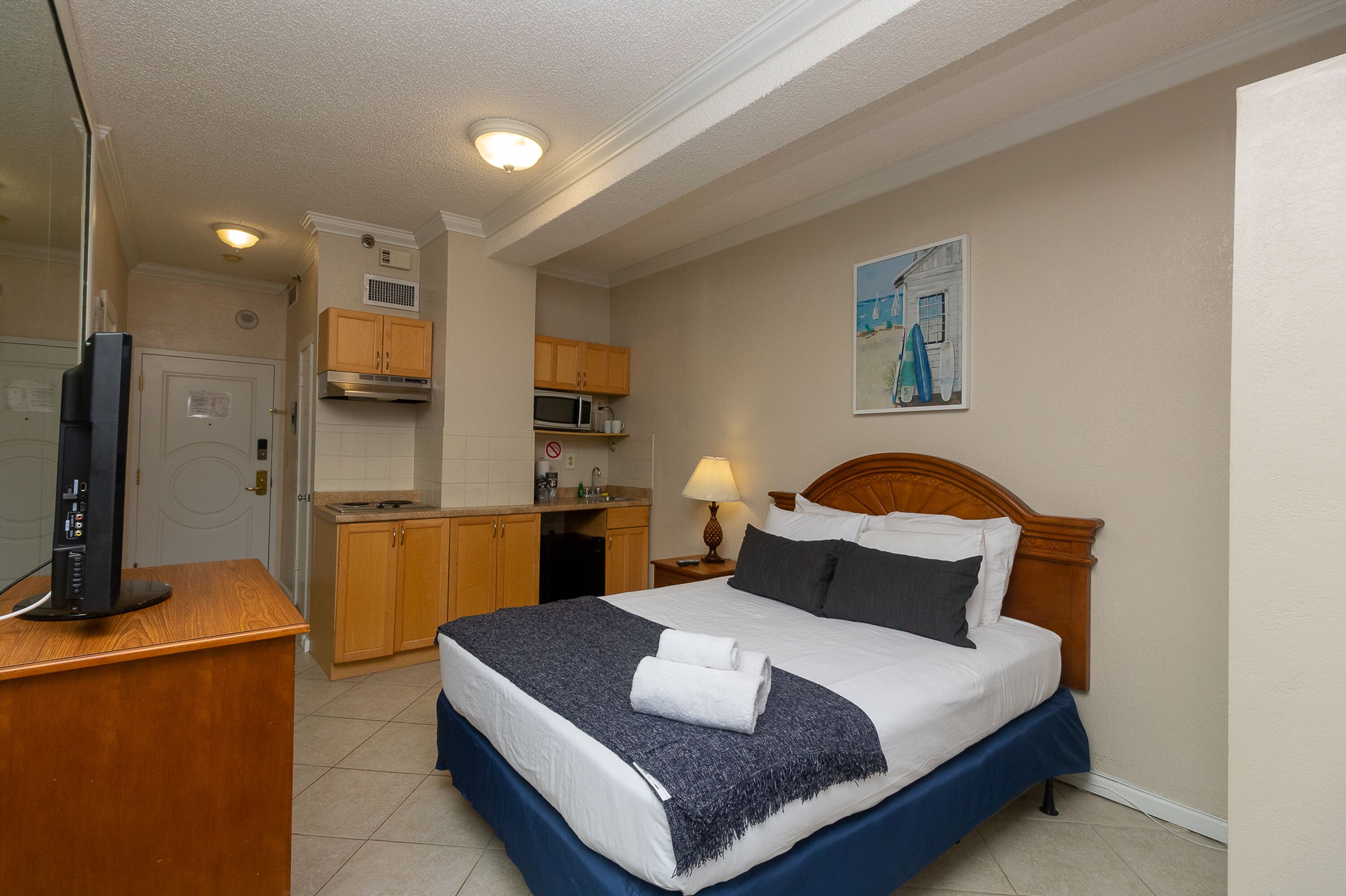Quarto de hotel com cama, escrivaninha, televisão, cozinha compacta e um quadro na parede.