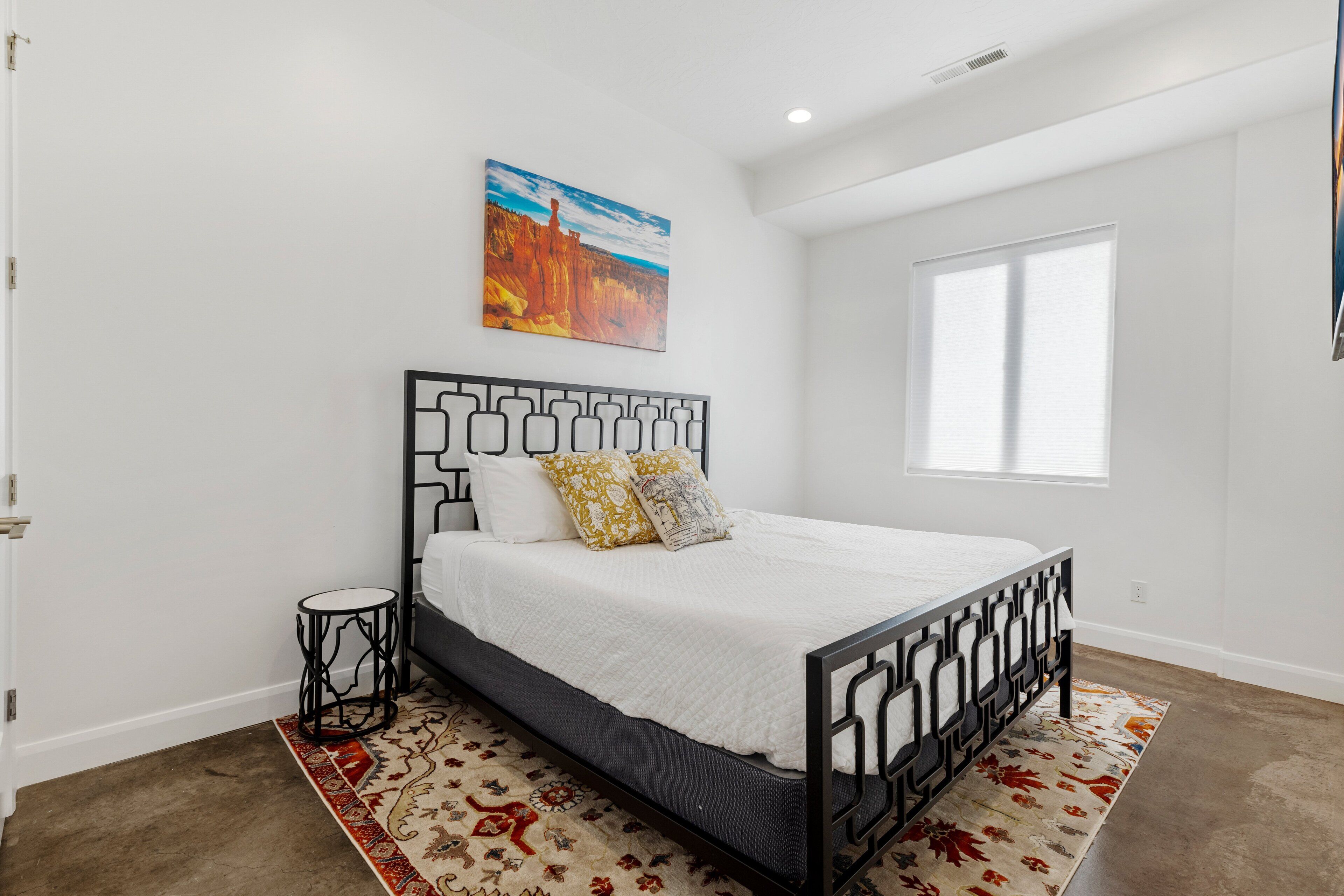 A bedroom with a bed, a small side table, a painting on the wall, and a window.