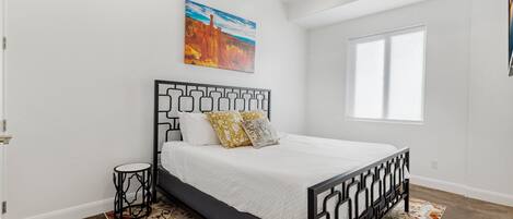 A bedroom with a bed, a small side table, a painting on the wall, and a window.