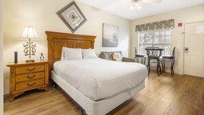Quarto com cama grande, mesa de cabeceira de madeira, abajur, ventilador de teto, quadro decorativo e janela com persianas.