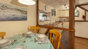 Ein Esszimmer mit Tisch, Geschirr, Gläsern und Besteck, Holzstühlen, einem Küstenmotiv als Gemälde, einer Uhr und einer Küchenzeile mit Schränken und Herd.