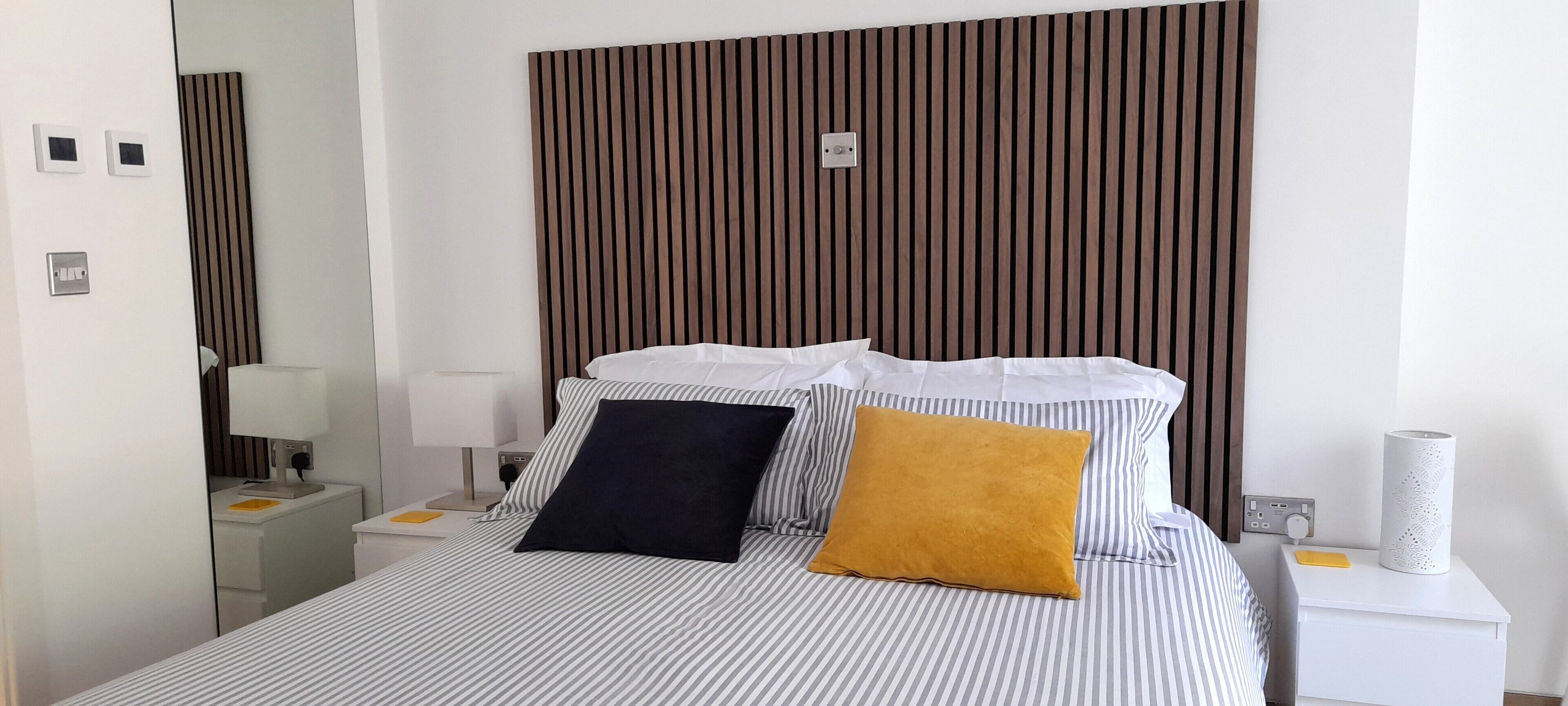 Quarto de hotel com cama listrada, cabeceira de madeira, mesa de cabeceira com abajur e um rolo de papel higiênico.