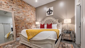 Une chambre avec un mur en briques, un lit avec une literie blanche et des oreillers rouges, un miroir et une table de chevet avec une lampe.