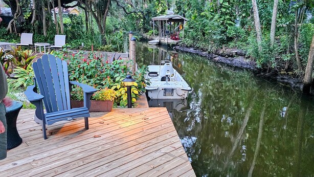 A wooden deck with a chair and potted plants, a small boat docked at a pier, and a gazebo in a lush, green environment.