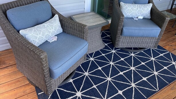 Twee rieten stoelen met blauwe kussens en geborduurde kussens, een klein rond tafeltje en een vloerkleed met een geometrisch patroon op een houten terras.