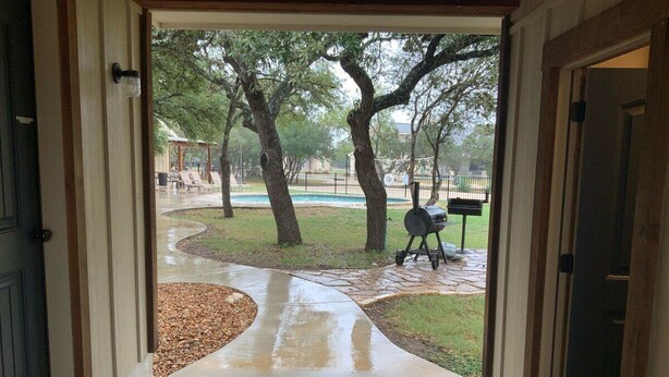 A wet concrete driveway leading to a covered patio area with a barbecue grill. - Double L River House @ Frio Premier Rental Homes (Concan)