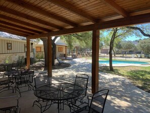 Um pátio ao ar livre com mesas e cadeiras de metal, uma piscina ao fundo e uma área coberta com vigas de madeira.